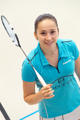 Jeune femme brune souriante, vêtue d'un tee-shirt bleue et arborant une raquette de badminton.